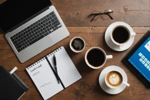 Composition à plat d'un ordinateur portable avec différents logos de médias sociaux sur son écran, une tasse de café et un bloc-notes avec une stratégie écrite pour acquérir des clients, sur une surface texturée en bois.